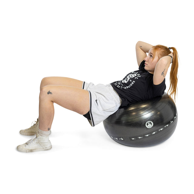 A person with long red hair works out by doing a sit-up on an Exercise Ball from Bells of Steel. They wear a black T-shirt and light shorts, highlighting their arm and leg tattoos, paired with white sneakers. The background is plain white.