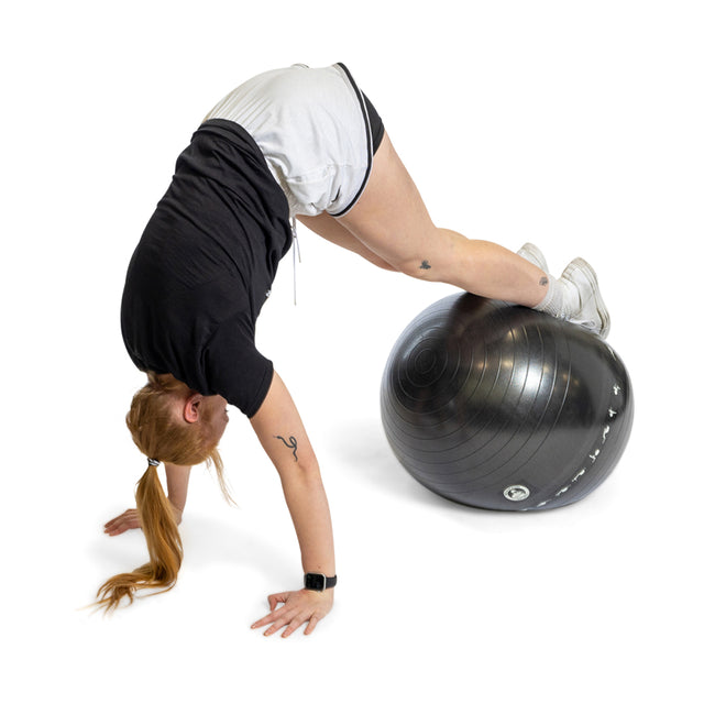 A person in a black shirt and white shorts performs a workout using the Bells of Steel Exercise Ball, balancing on it with their legs and hands on the floor to form an inverted V-shape.