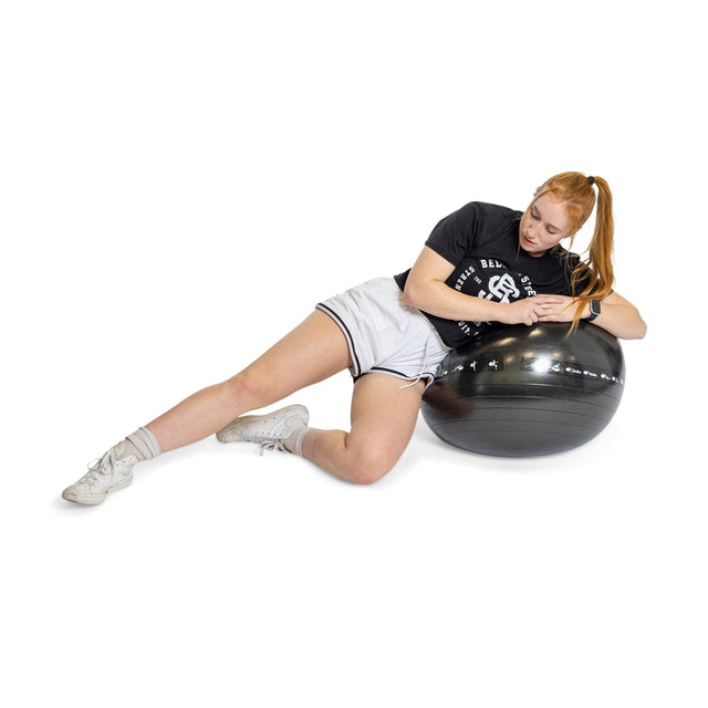 A red-haired woman in a black T-shirt and white shorts is resting on her side on an Exercise Ball from Bells of Steel. Set against a white background, she's wearing white sneakers and socks, skillfully balancing with her arms relaxed on the ball.