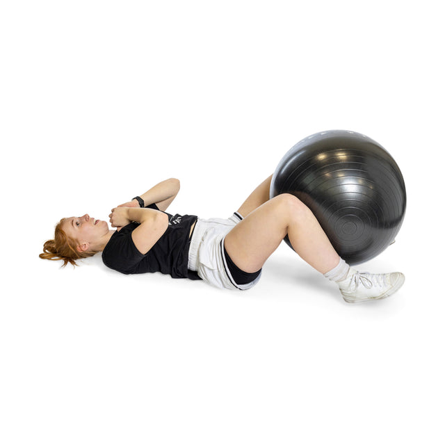 A red-haired person is lying on their back in a black shirt, gray shorts, and white socks with feet resting on a Bells of Steel Exercise Ball. Arms crossed over their chest against a plain white background.