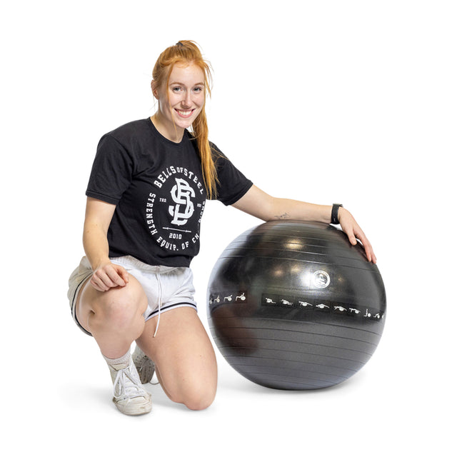 A person with long red hair kneels and smiles next to a large Bells of Steel Exercise Ball, ready for their workout in a black T-shirt, light gray shorts, white socks, and sneakers.