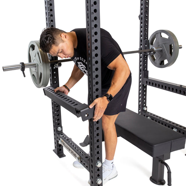 In the gym, a man dressed in a black T-shirt, shorts, and white shoes ensures training safety by adjusting the Flip-Down Safeties from Bells of Steel on a power rack with a loaded barbell.