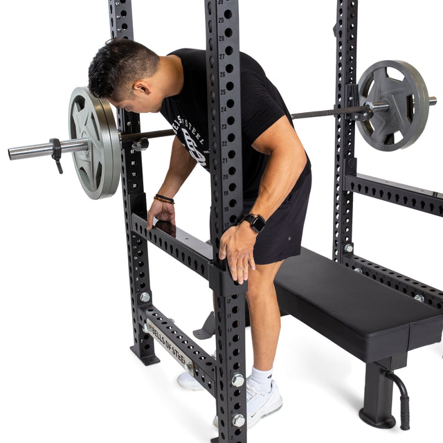 A person in a black shirt and shorts adjusts the Bells of Steel Flip-Down Safeties on a squat rack loaded with weights. The setup, which includes a bench, is placed against a white background. Focused on training safety, the individual diligently prepares for an intense workout session.