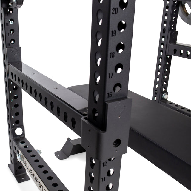 A close-up view showcases the Bells of Steel black adjustable power rack, designed with numbered holes. Featuring Flip-Down Safeties and a bench visible in the background, this squat rack prioritizes training safety with its sturdy metal construction.