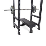 A Bells of Steel weight bench sits inside a black power rack with a loaded barbell on the supports. Flip-Down Safeties from Bells of Steel are installed for added training safety. The bench displays the "Bells of Steel" logo on its side.