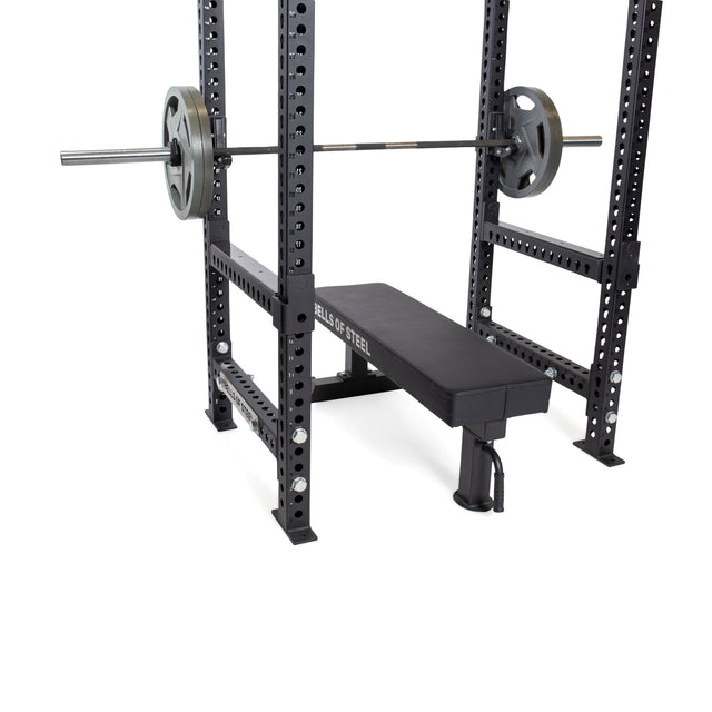A Bells of Steel weight bench sits inside a black power rack with a loaded barbell on the supports. Flip-Down Safeties from Bells of Steel are installed for added training safety. The bench displays the "Bells of Steel" logo on its side.