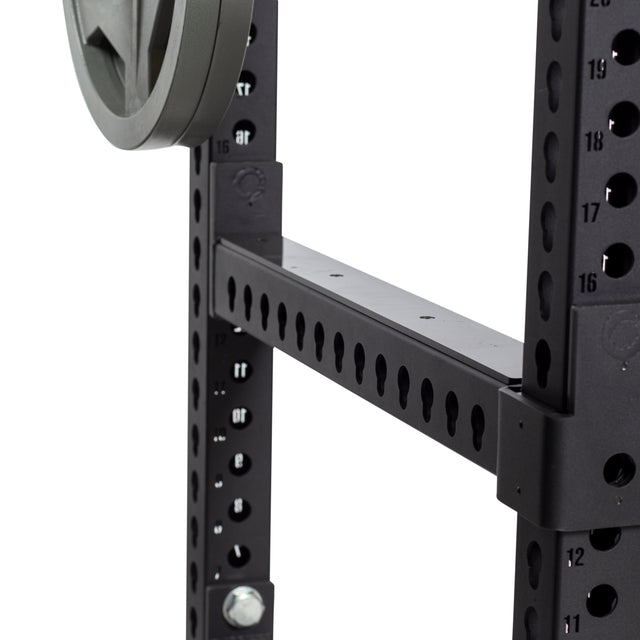 Close-up of a black adjustable crossmember on a power rack by Bells of Steel, showcasing multiple holes for customization and featuring Flip-Down Safeties for enhanced training safety. A weight plate is partially visible on the left, and the rack is marked with numbers for precise alignment.