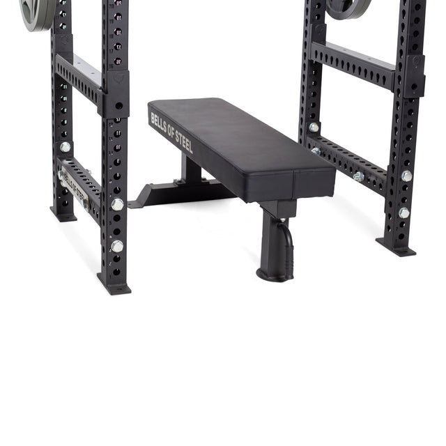 A close-up of a Bells of Steel weight bench inside a power rack featuring Flip-Down Safeties for enhanced training safety, with adjustable holes and attached weight plates.