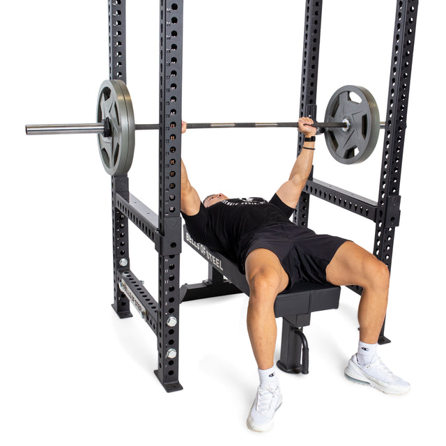 A person is performing a bench press on a black power rack, using the Bells of Steel Flip-Down Safeties to ensure enhanced training safety. They are lifting a barbell with weight plates on both sides, wearing a black T-shirt, shorts, and white sneakers while lying on a bench with arms extended upward.