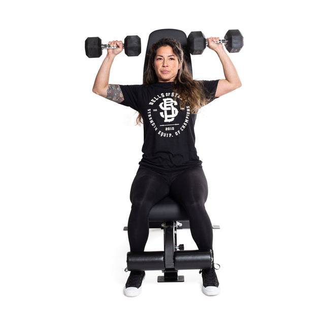 A person exercises on a Bells of Steel Flat/Incline/Decline Weight Bench, performing shoulder presses with dumbbells. They wear a black T-shirt with white text, black leggings, and black shoes. A plain white background emphasizes their focus on muscle targeting.