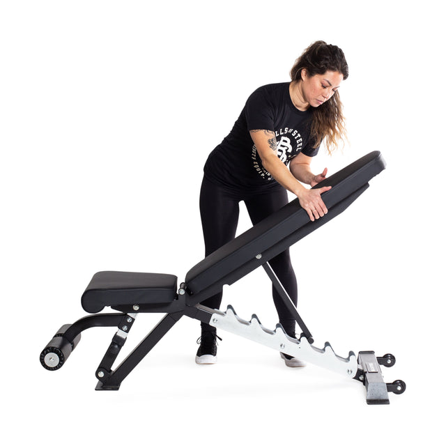 In a gym setting, a person adjusts the Bells of Steel Flat / Incline / Decline Weight Bench - Commercial, showcasing its adjustable nature and 1,000lb capacity. They are wearing a black t-shirt and leggings with sneakers. The bench features a padded seat and backrest, supported by a sturdy metal frame against a white background.