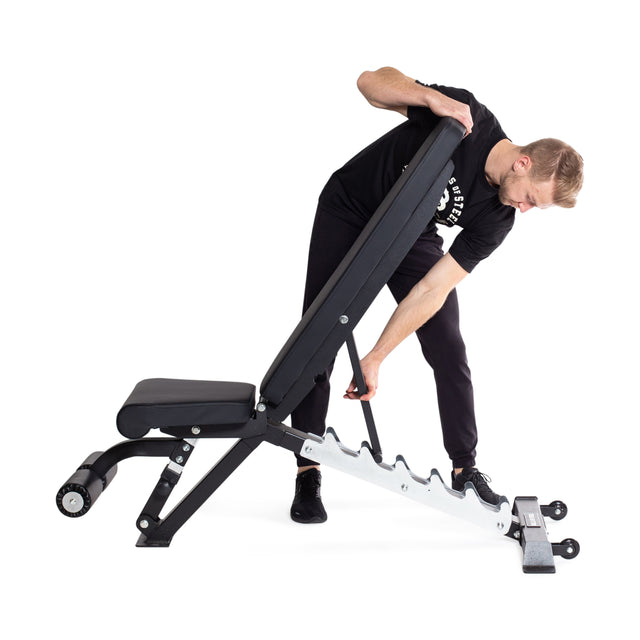 A person wearing a black shirt and pants adjusts the Bells of Steel Flat / Incline / Decline Weight Bench - Commercial, which has a 1,000lb capacity. This weight bench, designed for muscle targeting, features a padded seat and backrest with sturdy white metal supports set against a white background.