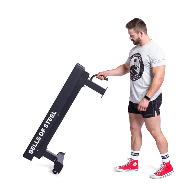 A person wearing a gray T-shirt, black shorts, and red sneakers effortlessly maneuvers a Bells of Steel Powerlifting Flat Bench. The gym equipment includes small wheels for easy movement and features an impressive maximum capacity.