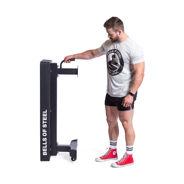 A man in a gray "Bells of Steel" t-shirt and red sneakers showcases how to move a folded gym equipment effortlessly using its wheels. The compact, black Powerlifting Flat Bench from Bells of Steel displays the brand's name on the side. It offers easy mobility and supports up to 1000lb, making it ideal for rigorous training sessions.