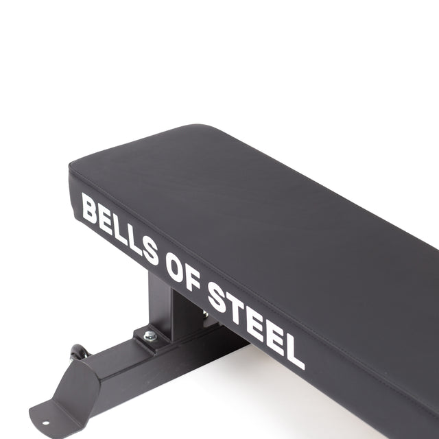 Close-up of a heavy-duty weight bench featuring the "Bells of Steel" brand name in white on its side. The Powerlifting Flat Bench showcases a simple, sturdy design with a black cushion.