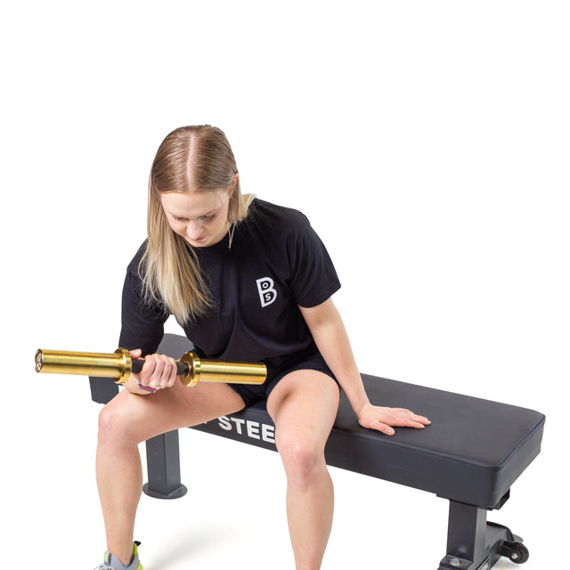 A person with long hair is seated on a Bells of Steel Powerlifting Flat Bench, holding a gold-colored barbell in one hand. They are wearing a black t-shirt, set against a plain white background.