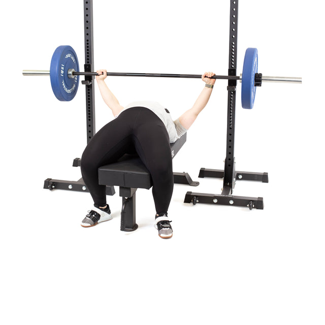 An individual is performing a bench press on the Bells of Steel Powerlifting Flat Bench, utilizing blue weight plates. They have a firm grip on the barbell positioned above their chest, supported by a 1,000lb max capacity setup in a power rack. The plain white backdrop of the gym accentuates their concentration and strength.