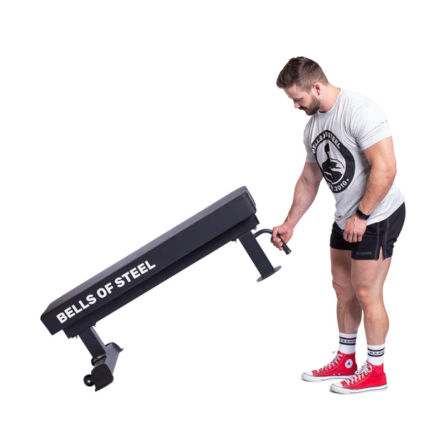 A person in a gray T-shirt and black shorts adjusts a Bells of Steel Powerlifting Flat Bench. They are wearing red high-top sneakers and standing against a plain white background.