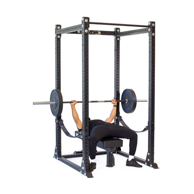 Inside a Bells of Steel power rack, a person performs a bench press on a flat bench, lifting a barbell. They wear black pants and Hydra Flat Foot Components shoes. The metallic rack includes multiple holes for easy adjustments.