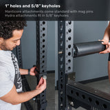 A man adjusts a bolt on the Bells of Steel Manticore Flat Foot Power Rack Builder (3" x 3", 1" Holes) while another person holds a cylindrical pad attachment. There are markings and numbers along the rack.