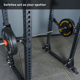 A barbell loaded with weight plates rests on the safety arms of the Bells of Steel Manticore Flat Foot Power Rack Builder (3" x 3", 1" Holes) in a gym. Text at the top reads, "Safeties act as your spotter.