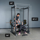 Two people in a gym use the Bells of Steel Manticore Flat Foot Power Rack - Prebuilt (3" x 3", 1" Holes) for bench pressing—one lifting while the other spots. Dotted lines outline the power rack’s dimensions: 49" wide, 54" deep, and 87¼" tall against a gray background, making it an ideal addition to your home gym setup.
