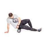 A man in a gray t-shirt and black pants uses the Bells of Steel Foam Roller under his right thigh, supporting himself with his left hand on the floor as he performs a self-myofascial release exercise.