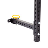 A close-up of a black metal bracket with 5/8" holes is shown attached to a vertical beam in a garage gym. This Bells of Steel Folding Rack 2.3" x 2.3" features a wall-mounted design and includes a horizontal bar with a yellow handle. The bracket is equipped with bolts and a locking pin, providing secure support for up to 600 pounds of static weight capacity.