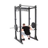 A man is squatting with a barbell inside a Manticore Flat Foot Components power rack from Bells of Steel. The black frame has numerous adjustment holes and the brand label. He's dressed in a black shirt, gray shorts, and black shoes.