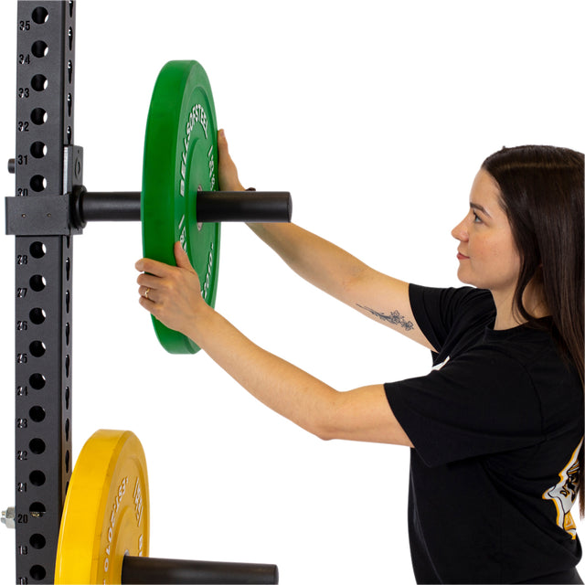 Wearing a black shirt, the individual with long brown hair and a tattooed arm effortlessly places a green weight plate onto the Bells of Steel power rack, where a yellow plate is already resting on the Pin Plate Pegs below. The rack showcases multiple holes for adjustable heights as they navigate the gym environment with ease.