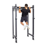 A person in a black shirt and shorts performs a pull-up on the Bells of Steel Fat/Skinny Pull Up Bar, which is built into a sturdy power rack with multiple adjustment holes.