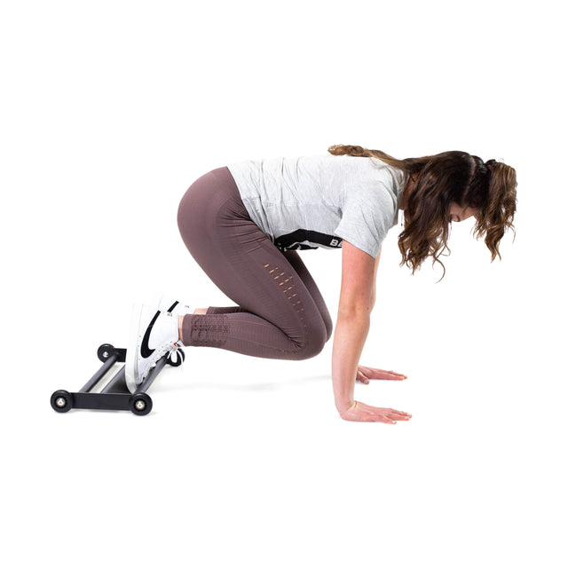 A person in athletic wear showcases an intense hamstring exercise using the space-efficient Glute Ham Slider by Bells of Steel. They are crouched with hands on the floor and feet on the slider, wearing a gray shirt, brown leggings, and white sneakers.