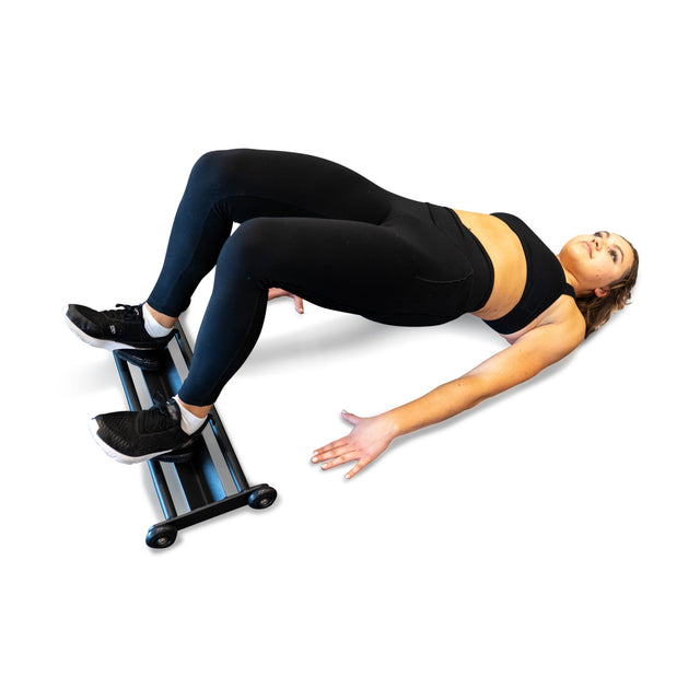 Dressed in workout attire, an individual uses the Bells of Steel Glute Ham Slider with foot rollers to perform a glute bridge. With knees bent at 90 degrees and arms extended for balance, this space-efficient design effectively targets the hamstrings.