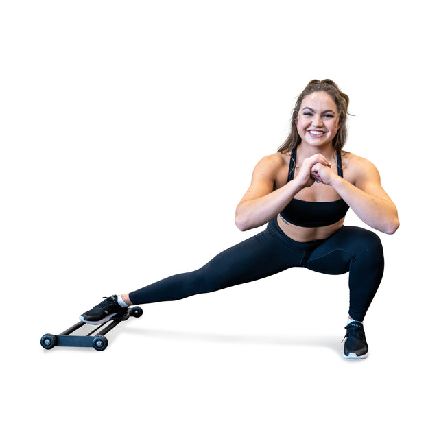 A woman in athletic wear performs a side lunge using the Bells of Steel Glute Ham Slider. Smiling with hands clasped at her chest and her right leg extended, the space-efficient design against a white background emphasizes her focused workout session.