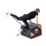 A woman in athletic wear uses the Bells of Steel Soft Glute Bench and a resistance band to perform a glute kickback, extending one leg backward while both hands rest on the bench for an effective home workout.