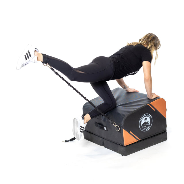 A woman in athletic wear uses the Bells of Steel Soft Glute Bench and a resistance band to perform a glute kickback, extending one leg backward while both hands rest on the bench for an effective home workout.