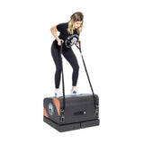 A woman works out at home on the Bells of Steel Soft Glute Bench, holding resistance bands attached to its sides. She wears black leggings, a black T-shirt, and white sneakers.
