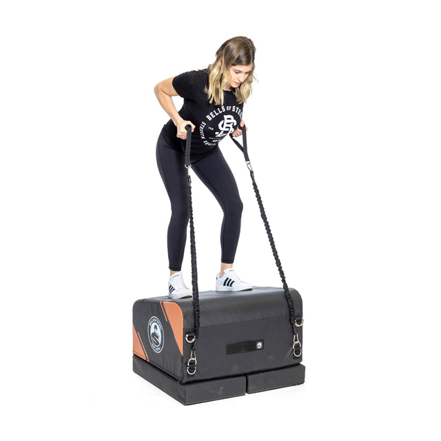 A woman works out at home on the Bells of Steel Soft Glute Bench, holding resistance bands attached to its sides. She wears black leggings, a black T-shirt, and white sneakers.