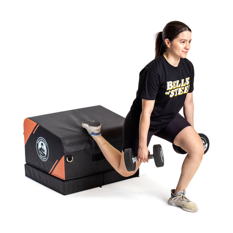 A woman performs a Bulgarian split squat with dumbbells, resting one foot on the Bells of Steel Soft Glute Bench. She is dressed in a Bells of Steel t-shirt, black shorts, and sneakers.