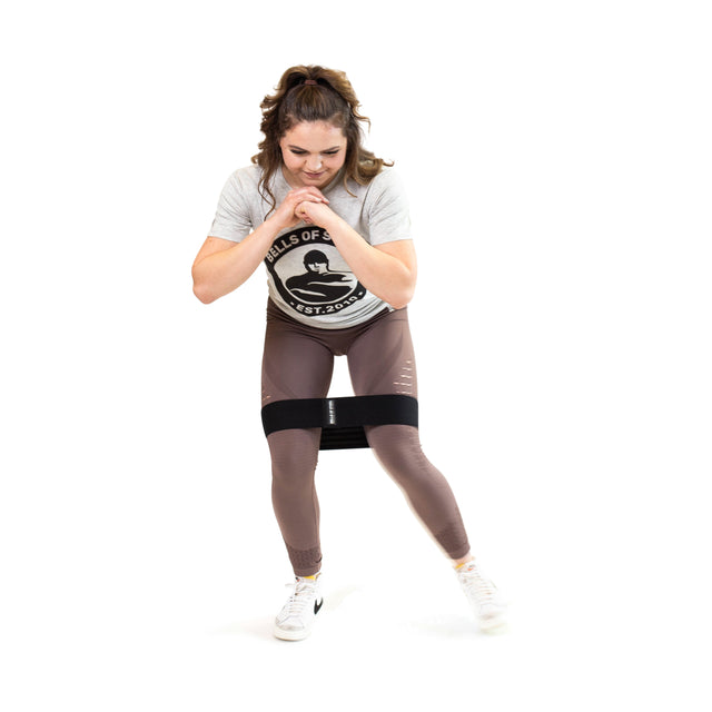 A woman is exercising with the Bells of Steel Glute Loop around her thighs, dressed in a grey t-shirt and brown leggings. The durable design of the band ensures it stays securely in place as she leans slightly forward with her hands clasped, all set against a white background.