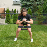 A woman demonstrates the Bells of Steel Ultimate Backyard Training Bundle, performing squats with a resistance band secured around her thighs. She trains outdoors on a grassy lawn, dressed in a black t-shirt, black shorts, and white sneakers. The backyard gym scene is complete with shrubs, a fence, and houses in the background.