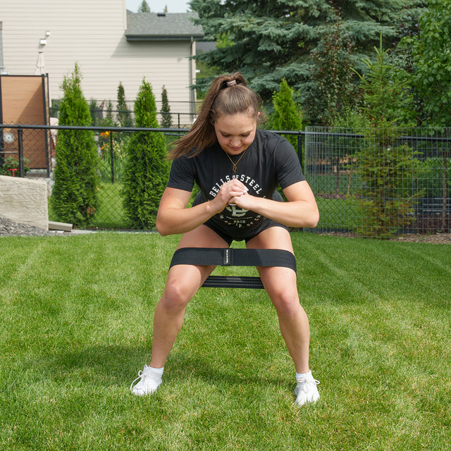 A woman squats on grass with a Bells of Steel Glute Loop above her knees, wearing a black shirt, black shorts, and white shoes—hands clasped for an effective booty workout.