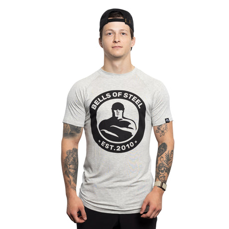 A young man wearing a backward black cap and a light gray Bells of Steel Bamboo T-Shirt stands facing forward, showing tattooed arms, against a plain white background.
