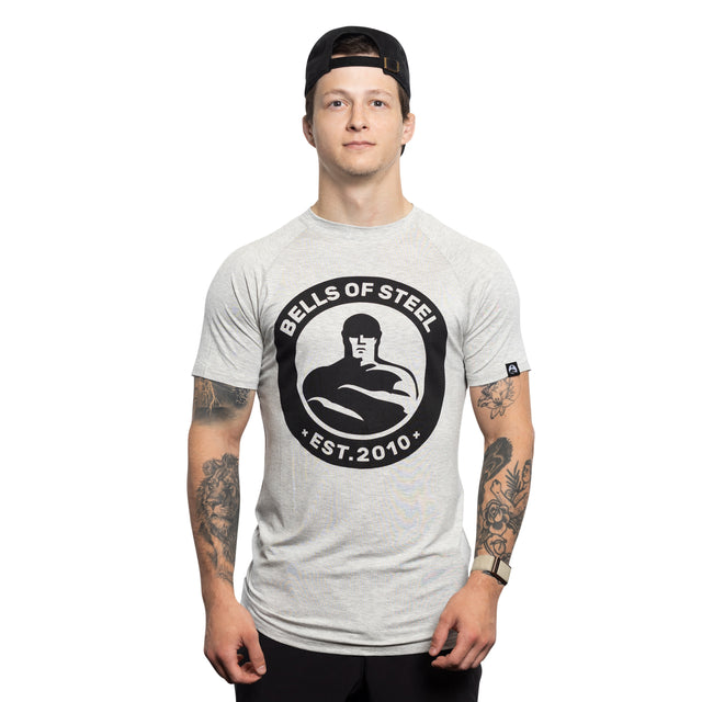 A young man wearing a backward black cap and a light gray Bells of Steel Bamboo T-Shirt stands facing forward, showing tattooed arms, against a plain white background.