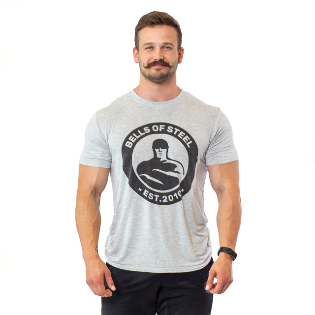 A man with a mustache stands against a white background, wearing a Bamboo T-Shirt from Bells of Steel, featuring a strongman figure. Complemented by black pants and a wristwatch, his attire provides comfort and sustainability.