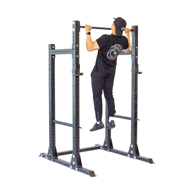 A person in black workout attire and a cap is performing a pull-up on the sleek Hydra Half Rack Builder by Bells of Steel, which includes 3" x 3" modular components with ⅝" holes for adjustable settings, all set against a crisp white background.