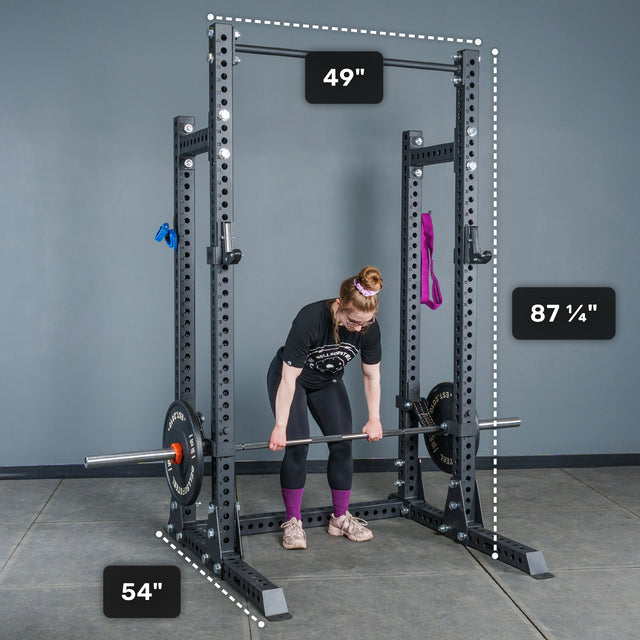 A woman prepares to lift a barbell inside the Bells of Steel Manticore Half Rack - Prebuilt (3" x 3", 1" Holes), a durable power rack ideal for any home gym, with labeled dimensions of 49" wide, 54" deep, and 87¼" tall. Resistance bands hang on the rack.