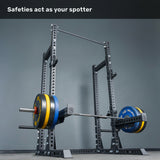 A loaded barbell rests on safeties within the Bells of Steel Manticore Half Rack - Prebuilt (3" x 3", 1" Holes). The text above reads, "Safeties act as your spotter." The background features a plain gray wall and floor—an ideal setup for any home gym.