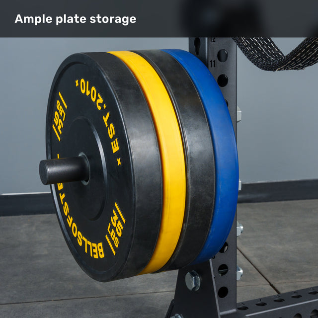 A close-up of the Bells of Steel Manticore Half Rack - Prebuilt (3" x 3", 1" Holes) showcases its sturdy uprights, holding several large black, yellow, and blue weight plates in a gym setting. The image features the text “Ample plate storage” at the top—making it a perfect addition to any home gym.