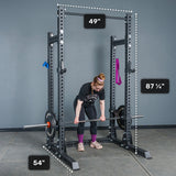 A person prepares to lift a barbell inside the Bells of Steel Manticore Half Rack - Prebuilt (3" x 3", 1" Holes), which features robust 3x3 uprights. The rack’s dimensions are clearly labeled: 49 inches wide, 54 inches deep, and standing at 87 ¼ inches tall—making it an ideal choice for any serious home gym setup. Purple and blue bands hang on the rack.